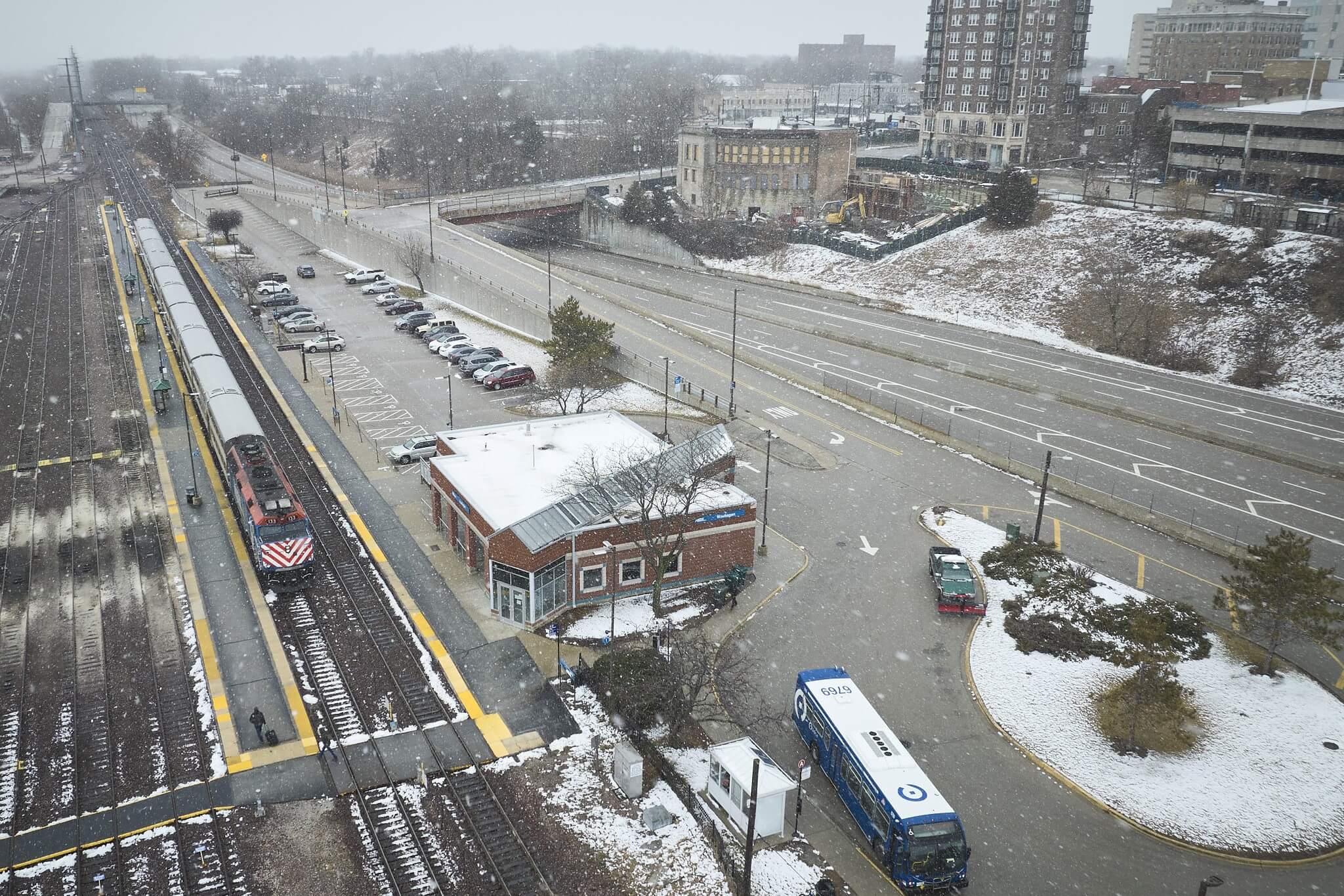 Aerial view of snow falling on train station. Metra train on tracks, Pace bus at bus stop. Truck with snow plow attachment.