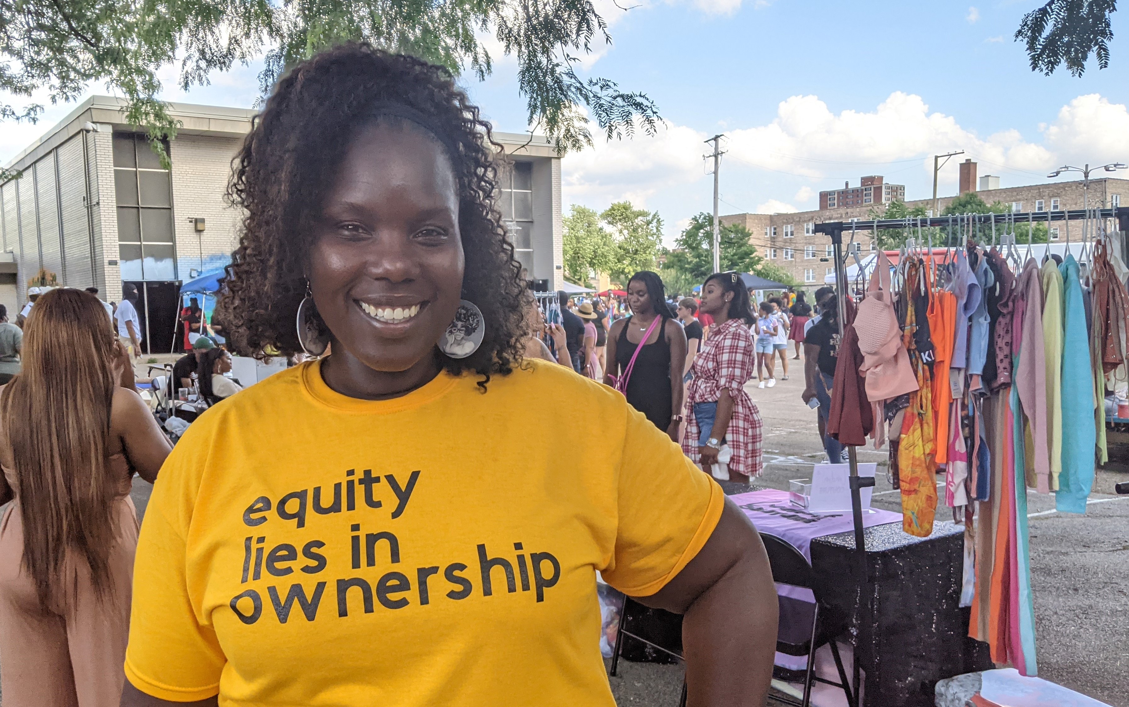 Smiling woman at outdoor festival with yellow t-shirt that says, "equity lies in ownership."